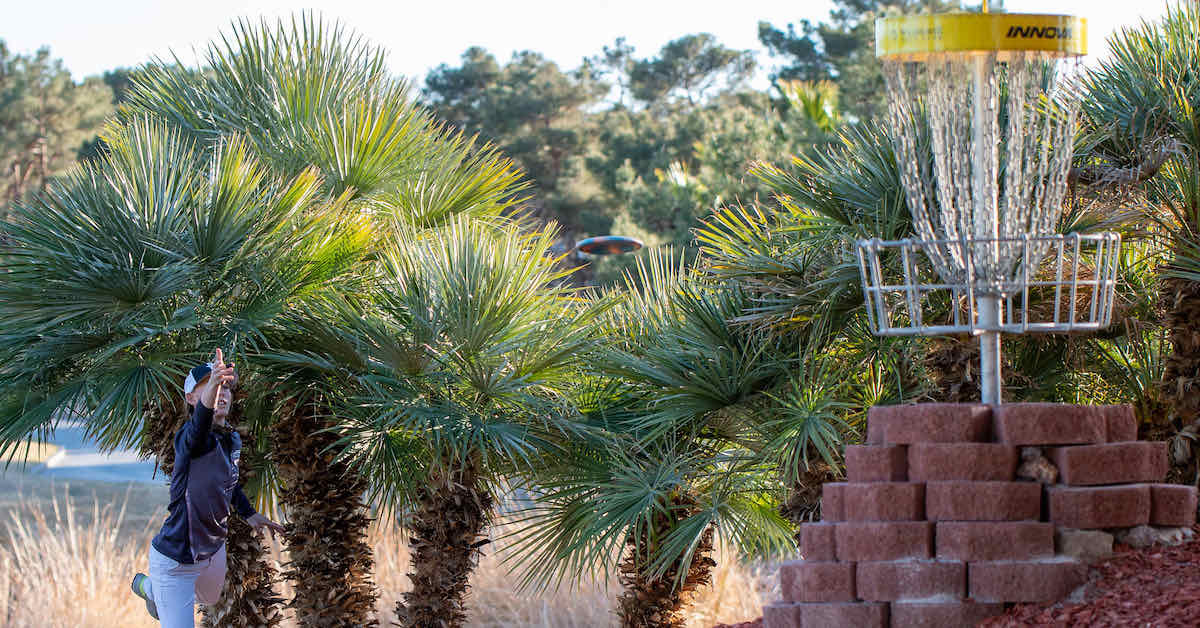 A woman putts at an elevated basket with palm trees nearby