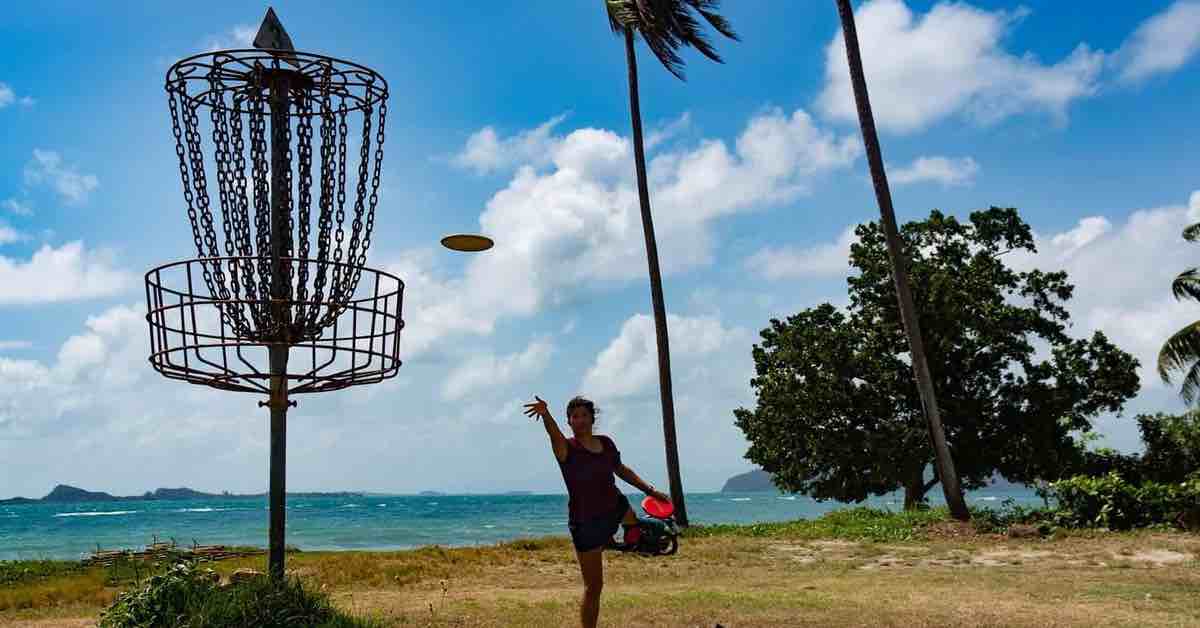 Woman putting to a disc golf basket on a tropical beach with ocean in distance