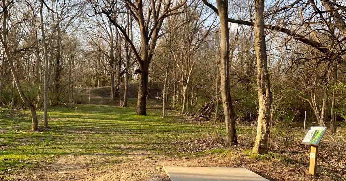 A concrete tee pad on a somewhat grassy disc golf fairway through a wooded area. Slight slope in the background of the picture