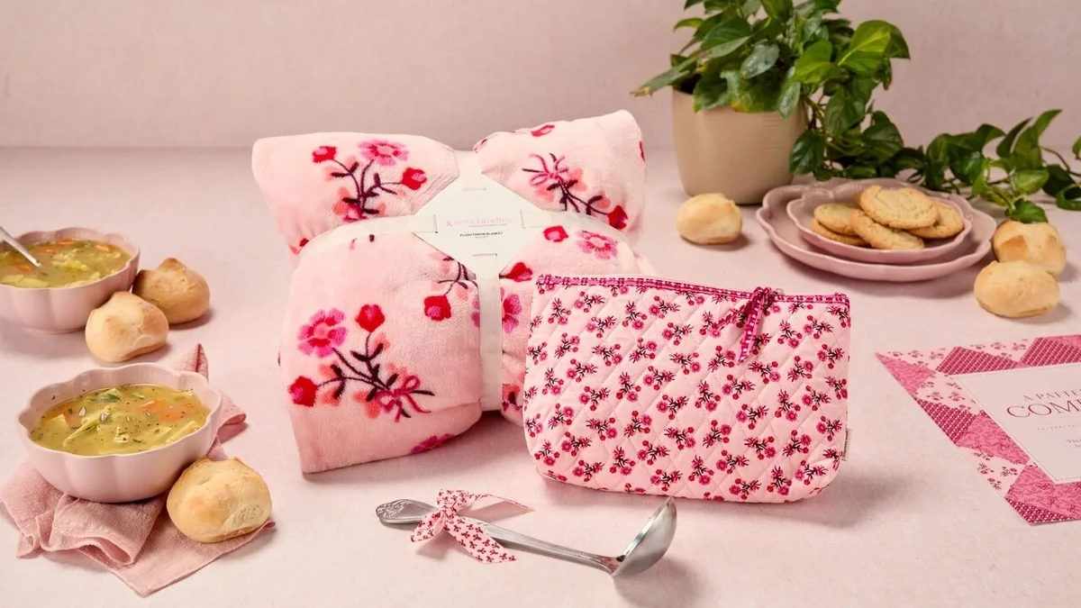 Pink-themed comfort gift arrangement featuring a soft floral throw blanket, matching floral pouch, two bowls of soup, rolls, cookies, and a metal ladle tied with a pink patterned ribbon.