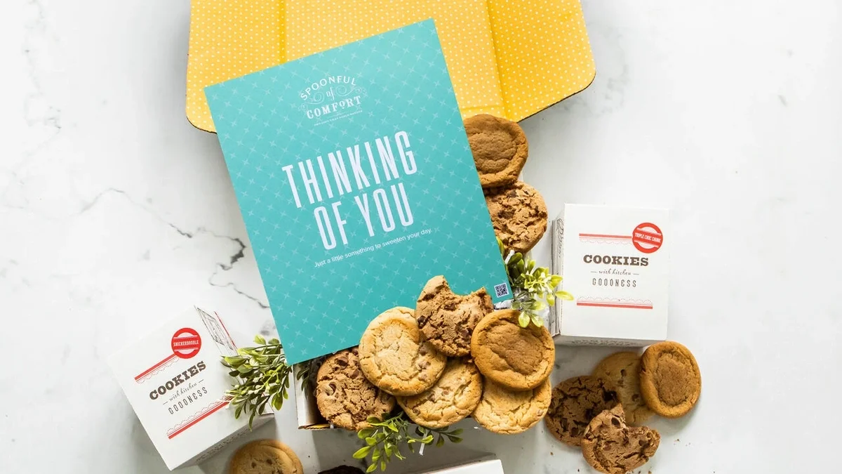 A Spoonful of Comfort Cookie package sits open on a white table, with a dozen varied cookies and a blue card that reads, "Thinking of You"