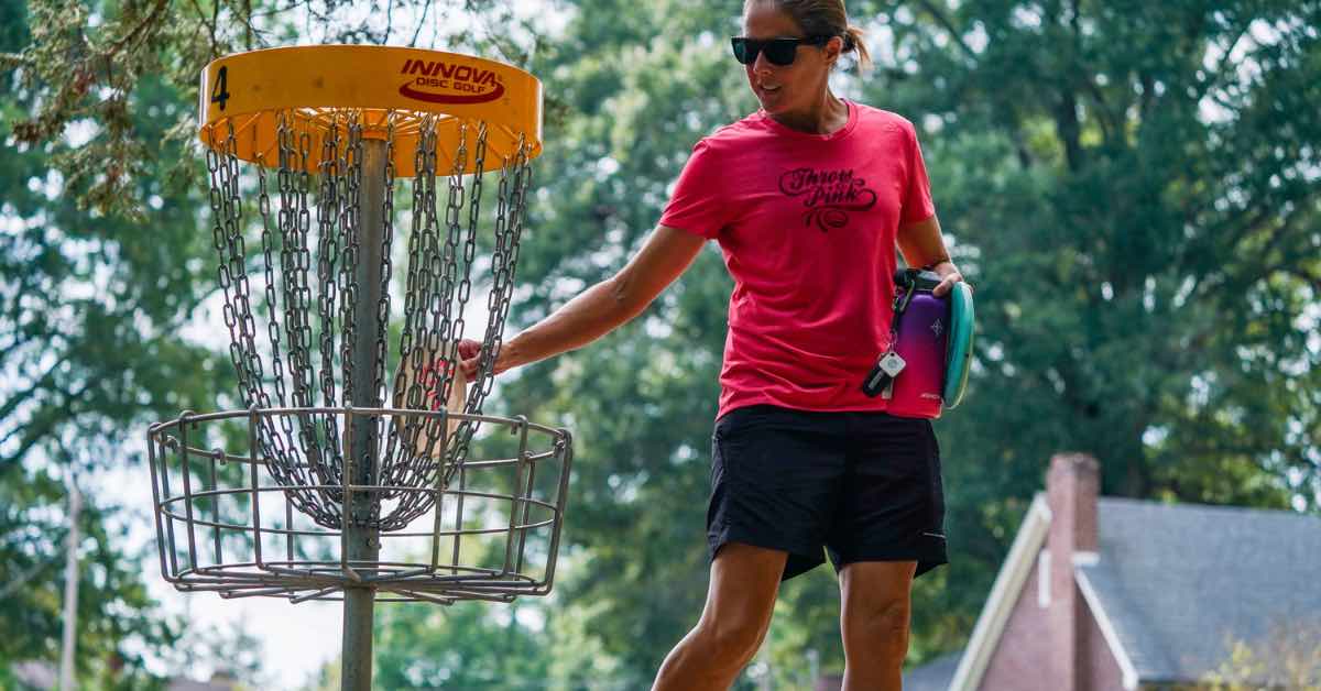 Woman in a pink shirt picks up a disc out of a disc golf basket
