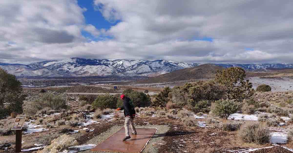 A man throwing from a disc golf tee pad with majestic mountains in the distance