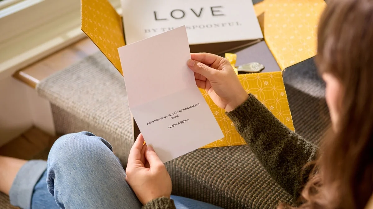 Person sitting with an open Spoonful of Comfort gift box, reading a heartfelt greeting card, with a cozy throw blanket and a card that reads “Love by the Spoonful” in the background.