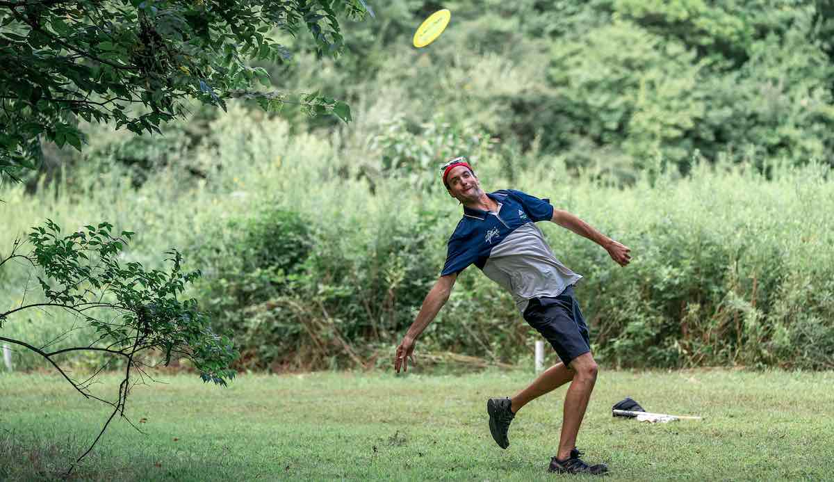 A man is in an awkward leaning position after throwing a disc golf shot on a grassy fairway