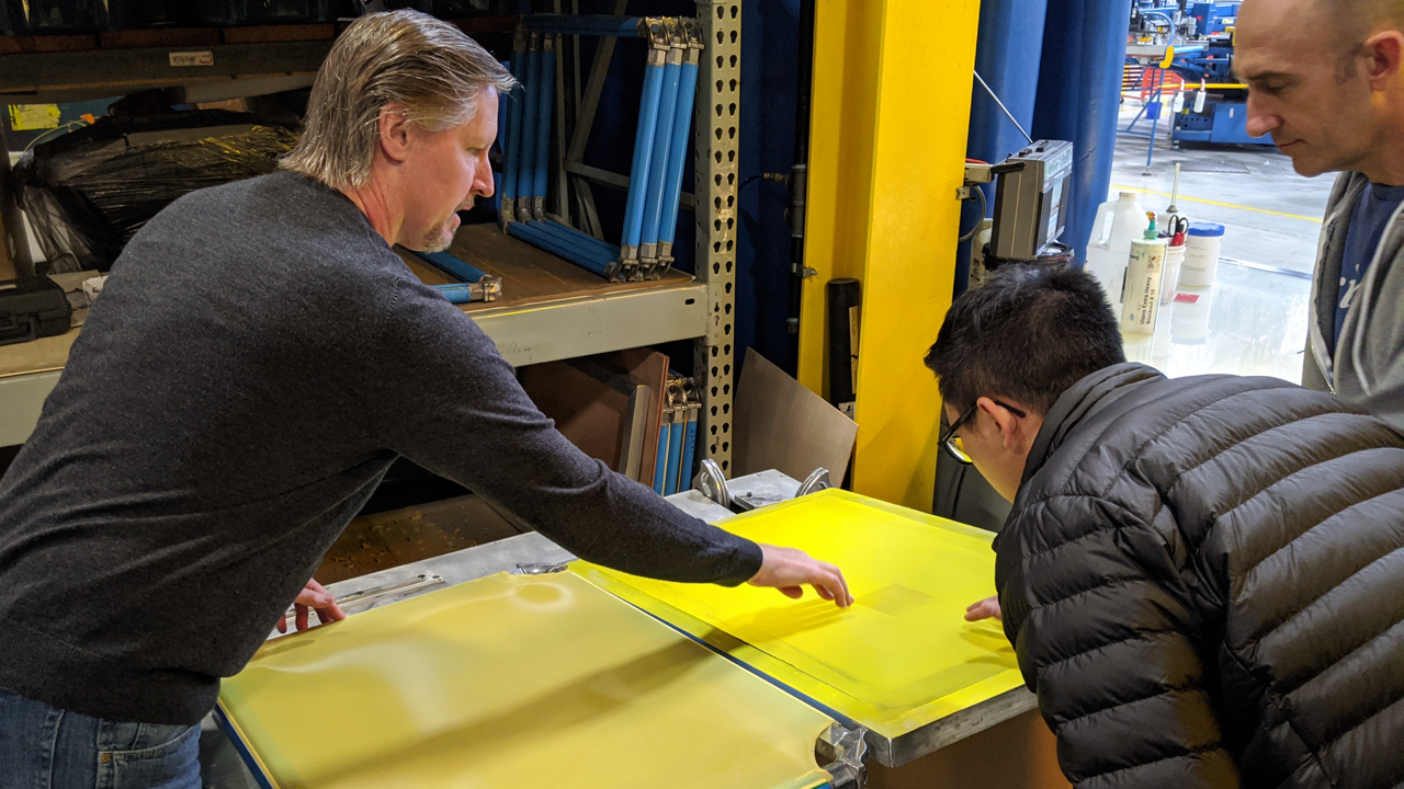 Two people closely inspect a thin-mesh screen printing screen at The M&R Companies.