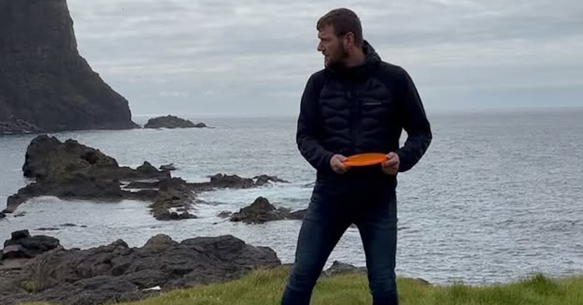 A man holds a disc golf disc in front of a sea and cliff backdrop