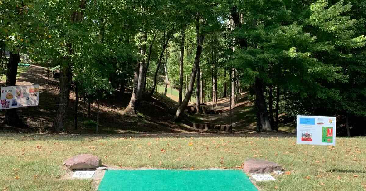 A turf disc golf tee in an open area leading to a wooded fairway between hills