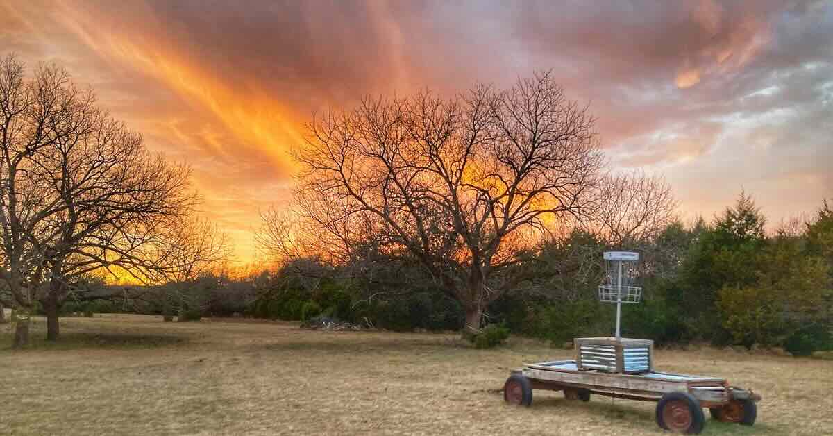 A disc golf basket on an old trailer in a field surrounded by trees at sunset