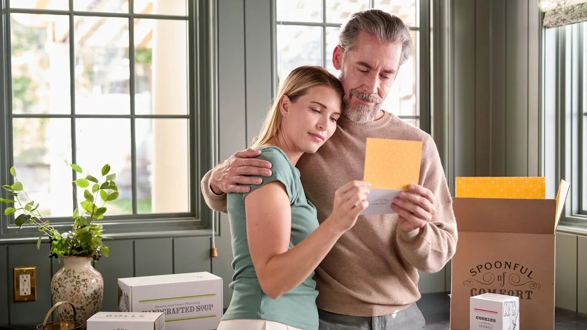 Couple standing together in a bright room, embracing while reading a greeting card beside an open Spoonful of Comfort gift box and packaged soup and cookies.