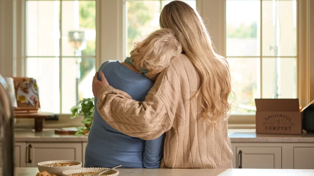 Two people embracing in a bright kitchen, with one resting their head on the other's shoulder. Bowls of soup sit on the counter, and a Spoonful of Comfort gift box is visible in the background.
