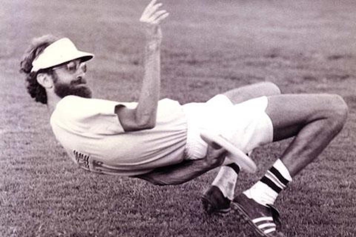 A black and white photo of a tall, lanky man catching a frisbee from behind his backa position nearly laying on the ground