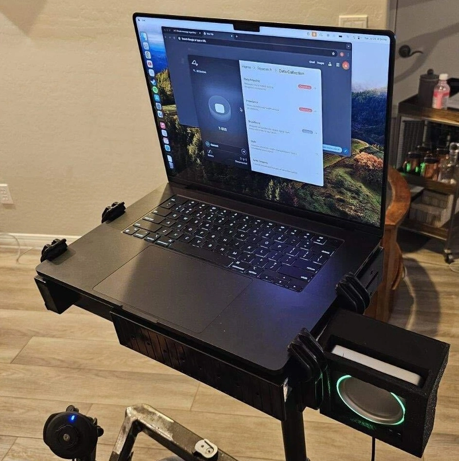 A Mac laptop photographed from the vantage point of a wheelchair. The laptop is on a stand attached to the wheelchair and attached to the right side of the laptop is a black plastic box with an inset circular metallic object surrounded by a glowing ring.