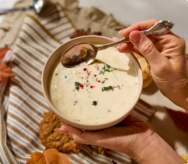 Potato Soup being held by a hand and spoon dipping into the sauce, a gingham towel is displayed in the backgorund