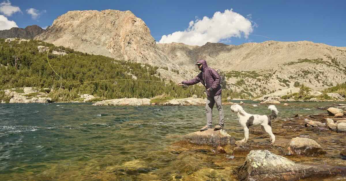 A man casting for fly fishing into a river in a remote mountain landscape as his dog looks on