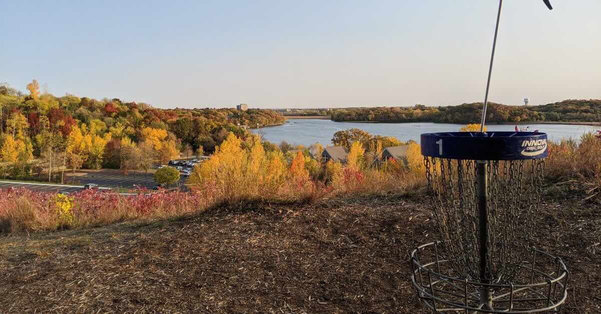 A blue disc golf basket in an elevated area with a view of a lake and a landscape in fall colors