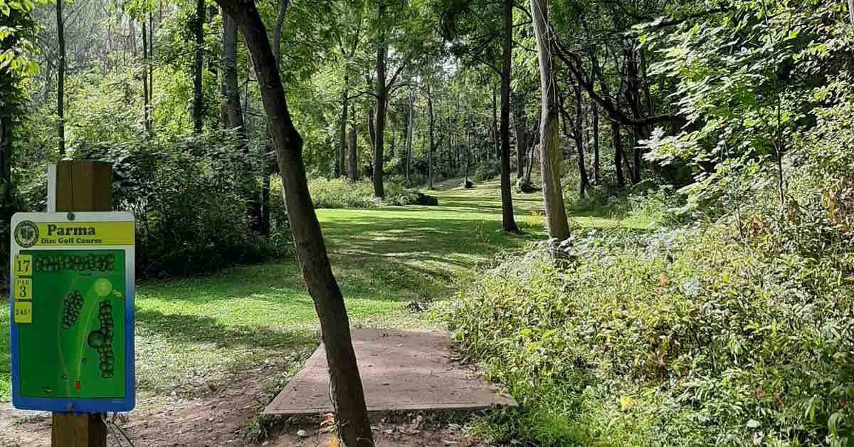A concrete disc golf tee pad leads to a wooded fairway. Parma Disc Golf tee sign in foreground