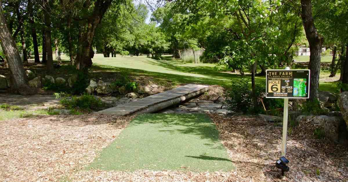 A turf disc golf tee pad on wood chips with bridge andscattered trees