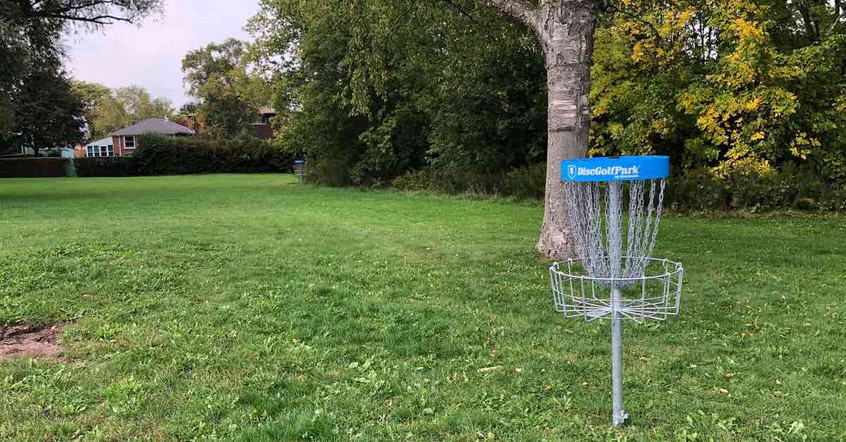 A blue-banded disc golf basket in a park setting with suburban houses in the background