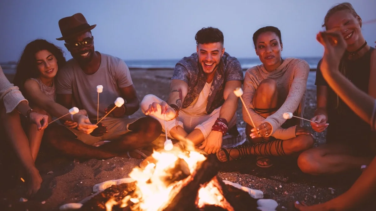 Group of friends gathered around a campfire at dusk, smiling and roasting marshmallows together.