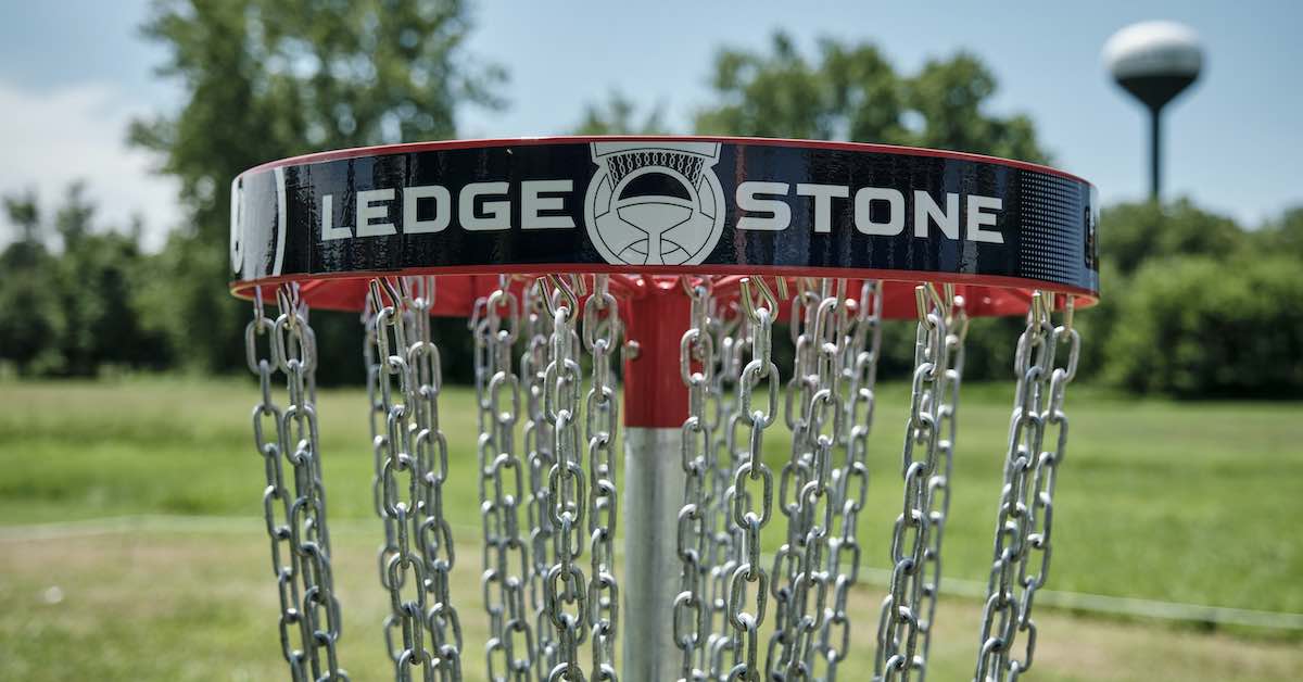 Top of a disc golf basket that says "Ledgestone." A water tower is scene blurred in the far background