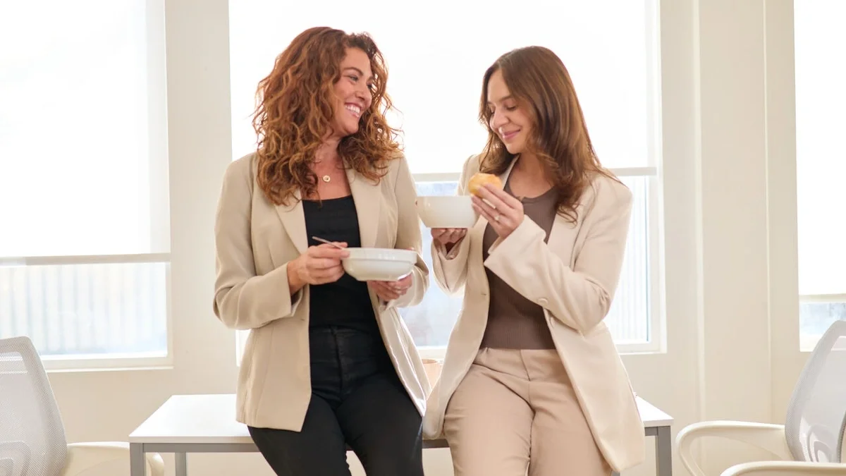 Two coworkers standing together in a bright office, smiling and enjoying bowls of soup during a shared meal.