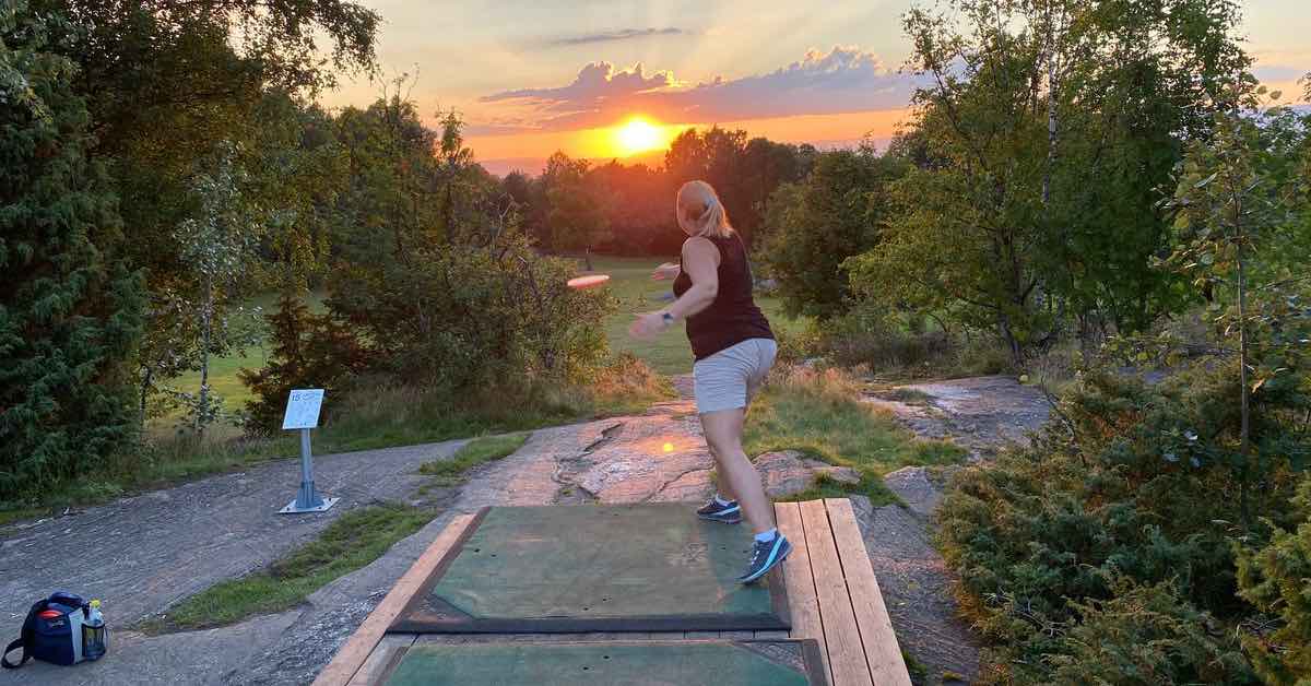 A woman throws a disc from a tee pad on a rock into the setting sun