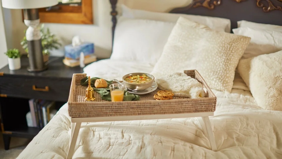 Food tray on a bed holding a bowl of chicken soup, a roll, cookies, juice, and a gold bedside bell, surrounded by cozy pillows and blankets in a bedroom.