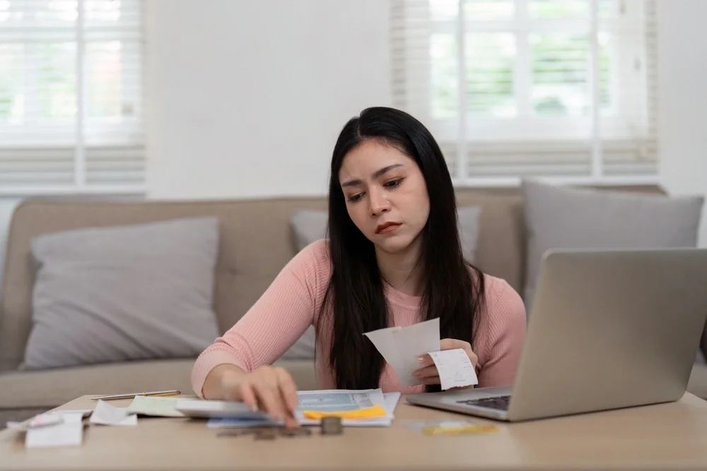 young woman sat at her dining table looking through documents and receipts with her laptop open