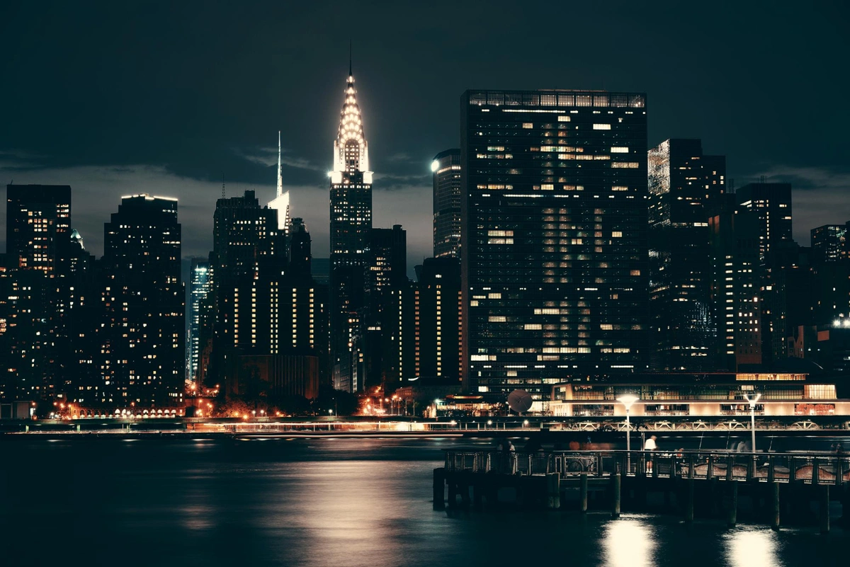 A nighttime cityscape featuring illuminated skyscrapers, with the iconic Chrysler Building prominently glowing in the center. Reflections from the buildings shimmer on the calm water in the foreground, alongside a pier with soft lighting.
