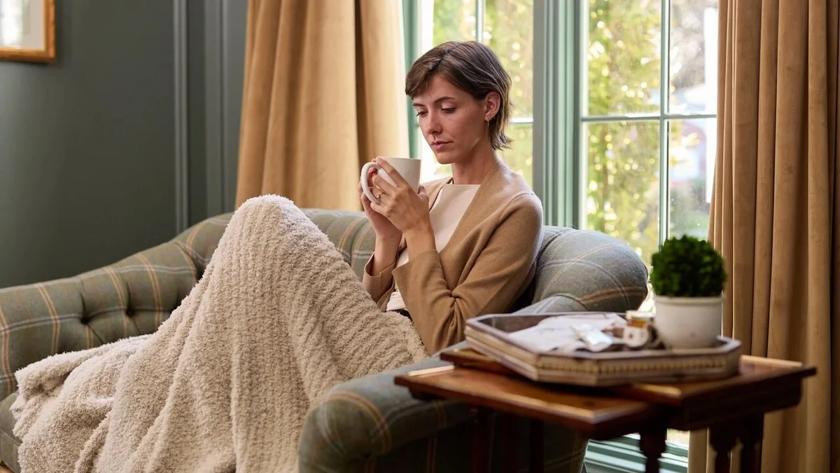 Person relaxing in an armchair under a cozy blanket, holding a warm mug with both hands beside a sunlit window.