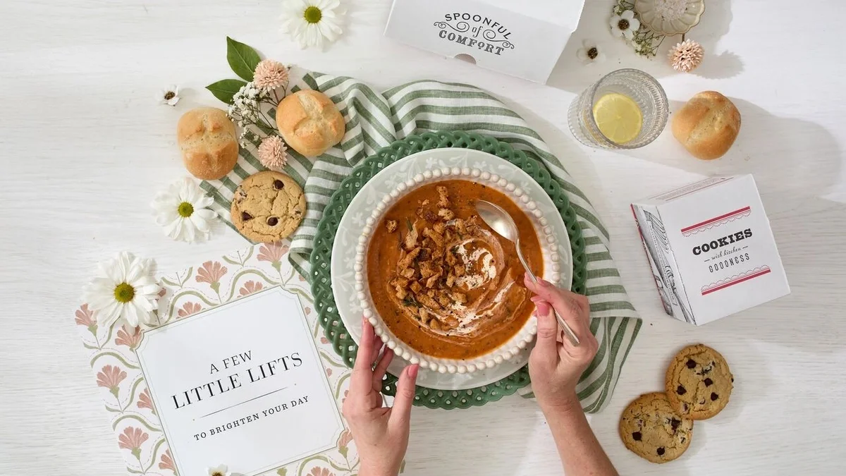 Overhead view of a styled tabletop featuring a bowl of creamy soup topped with a swirl of cream and crunchy garnish, held by a person with a spoon. Surrounding the bowl are cookies, bread rolls, a lemon slice in a glass dish, flowers, and branded cards reading “A Few Little Lifts to Brighten Your Day” and “Cookies,” creating a warm comfort-food presentation.