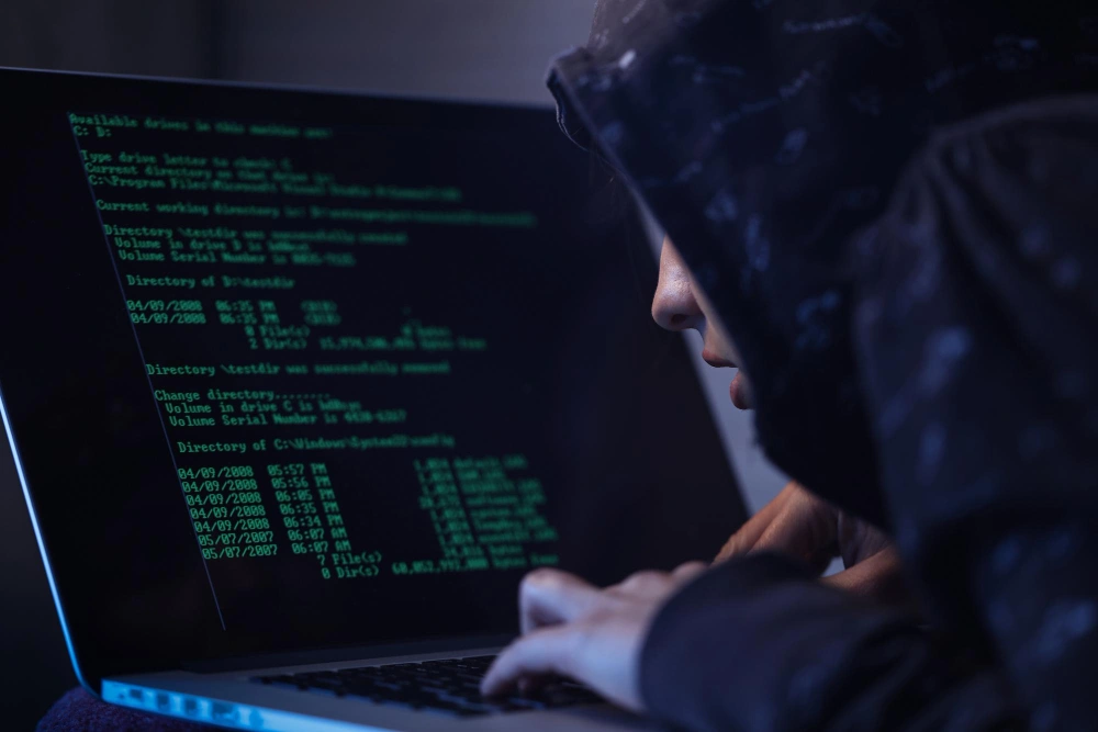 A person wearing a hoodie closely examining a laptop screen displaying code, symbolizing cybersecurity threats or hacking activities in progress.