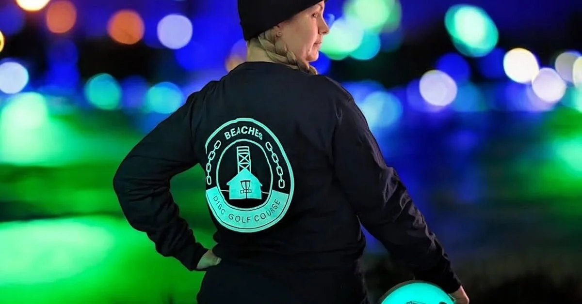 A woman in focus in front of a blurry background of lights while she hold a glow disc golf disc