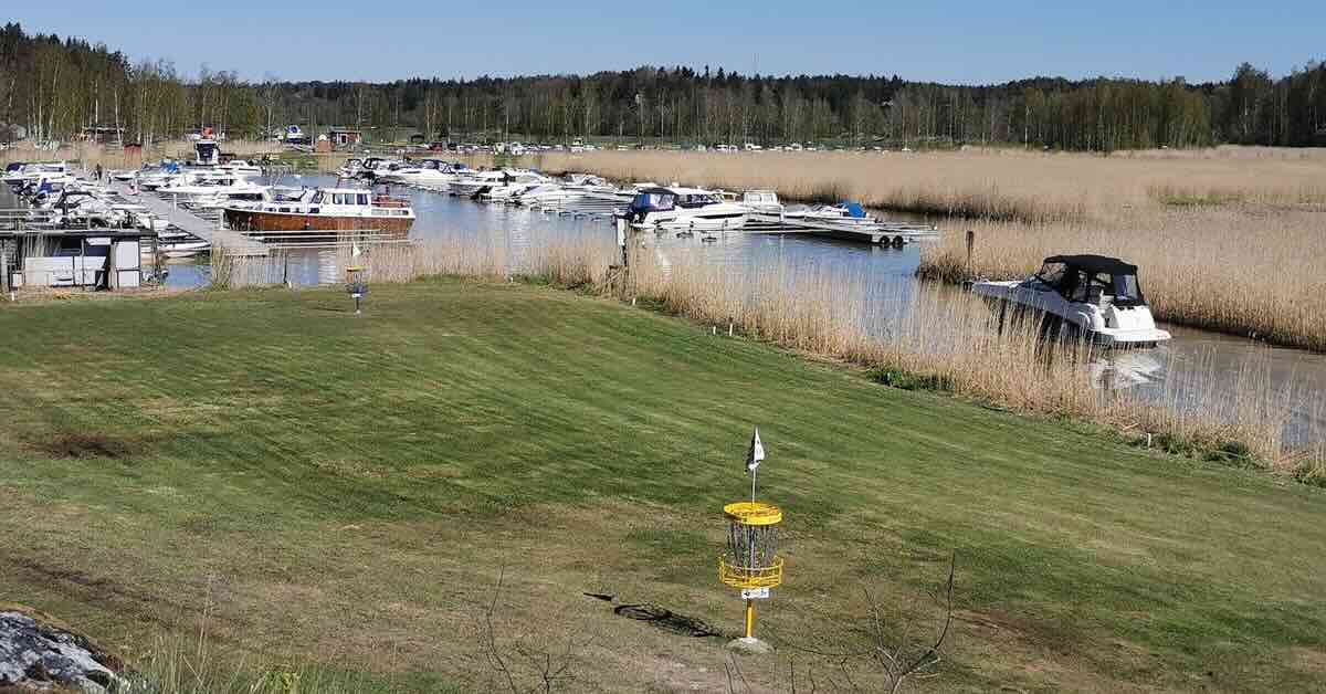 A yellow disc golf basket on flat, well-mown ground near a harbor full of boats