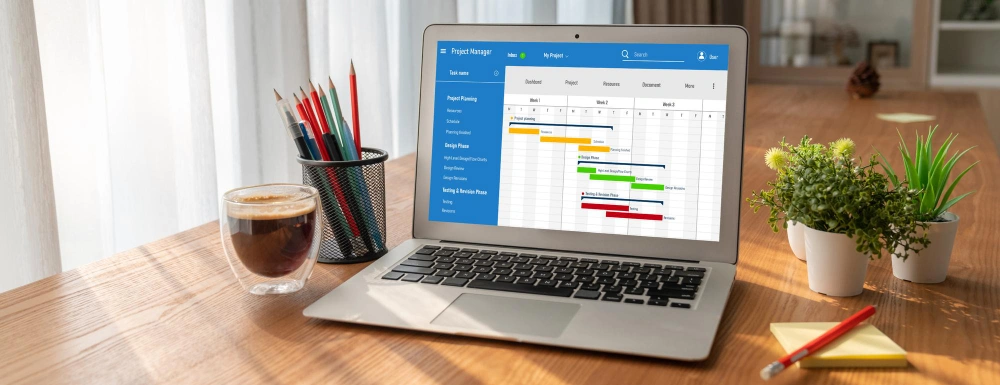 A laptop on a wooden desk displaying a Gantt chart for project management, with a cup of coffee and stationery on the side.