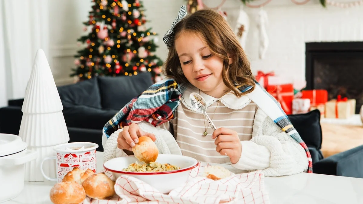 Young child wrapped in a plaid blanket, enjoying a bowl of soup with fresh rolls at a table, with a decorated Christmas tree and presents in the background.