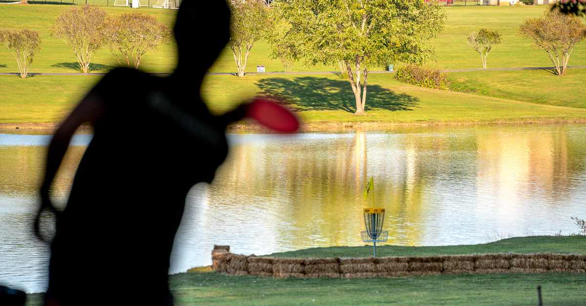 Silhouette of a player holding a disc. They are elevated over a basket below that is just in front of a lake