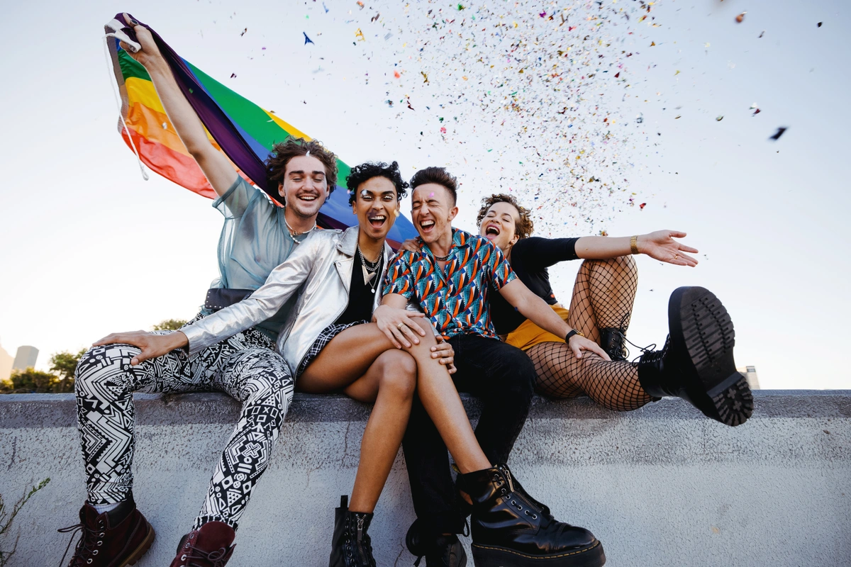 A group of queer folks laughing with a pride flag celebrating LGBTQ joy.