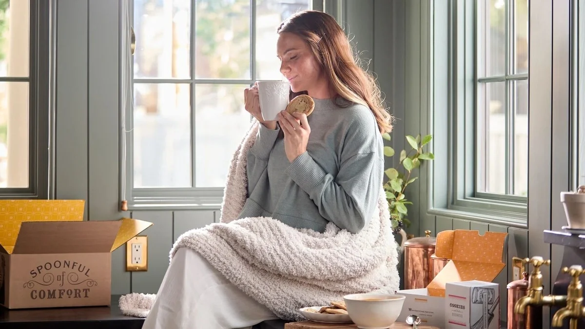 Woman wrapped in a cozy throw blanket, sitting by a bright window while enjoying a warm drink and a cookie, with open Spoonful of Comfort gift boxes and treats nearby.