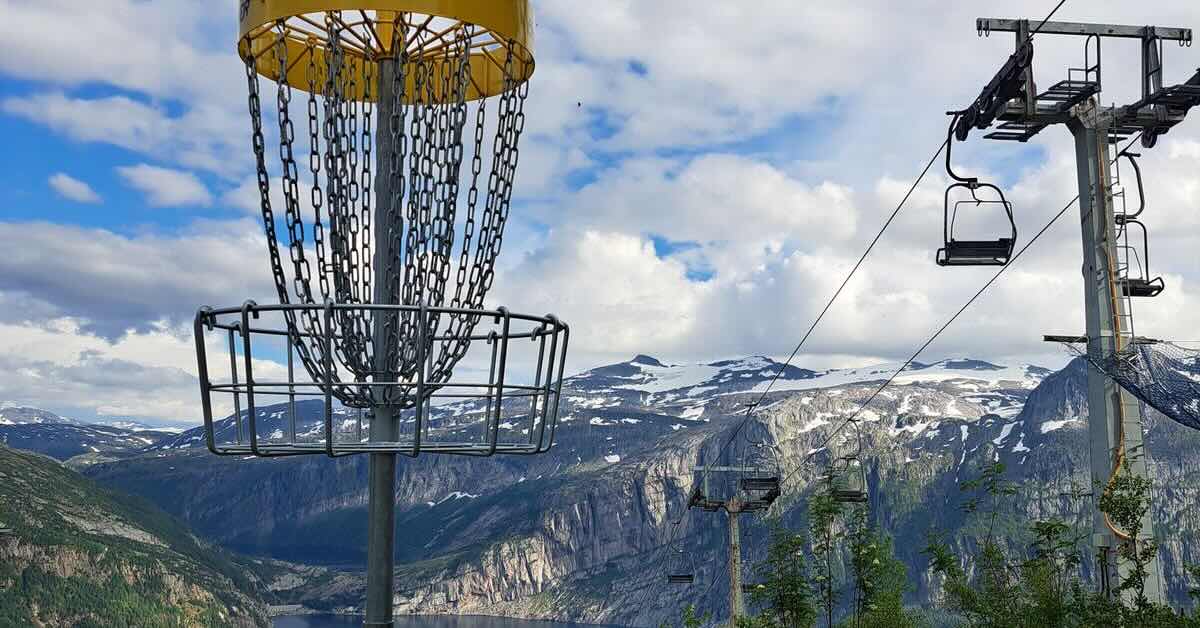 A disc golf basket with a backdrop of snow-capped mountains, water, and a ski lift