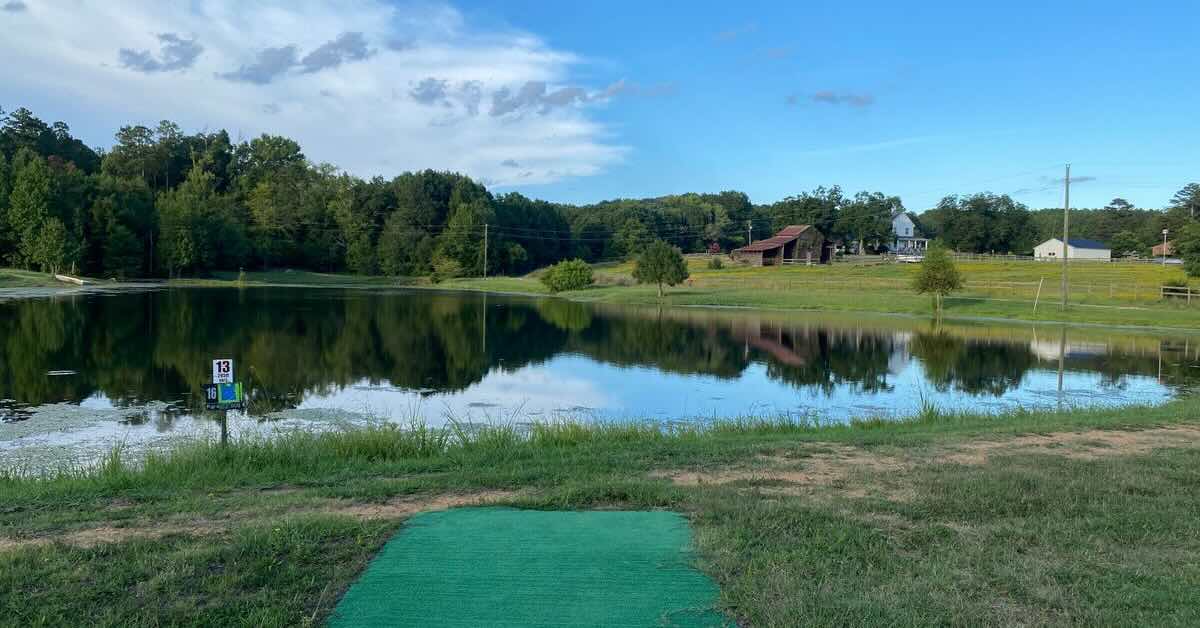 A turf tee pad in front of a large pond and an old farmhouse in the distance