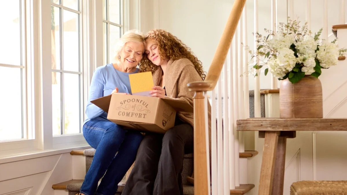 Two women sitting closely together on a staircase, smiling and embracing while holding an open Spoonful of Comfort gift box.