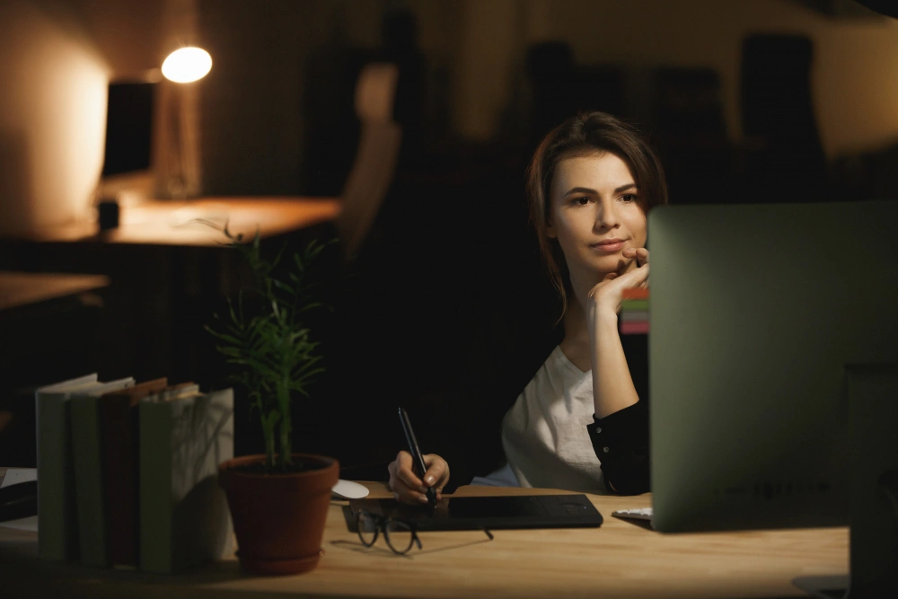 A young woman working late at night, sitting at her desk and looking thoughtfully at her computer screen.