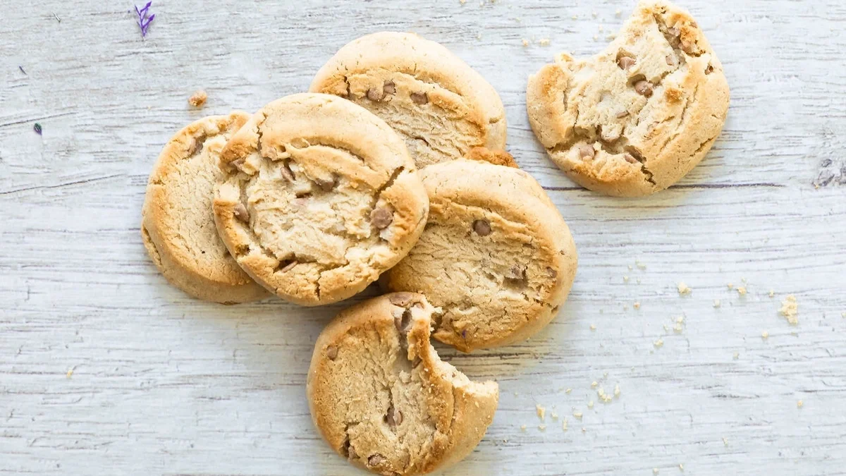 Close-up of five cookies on a light wooden surface, with one cookie partially eaten and scattered crumbs nearby.
