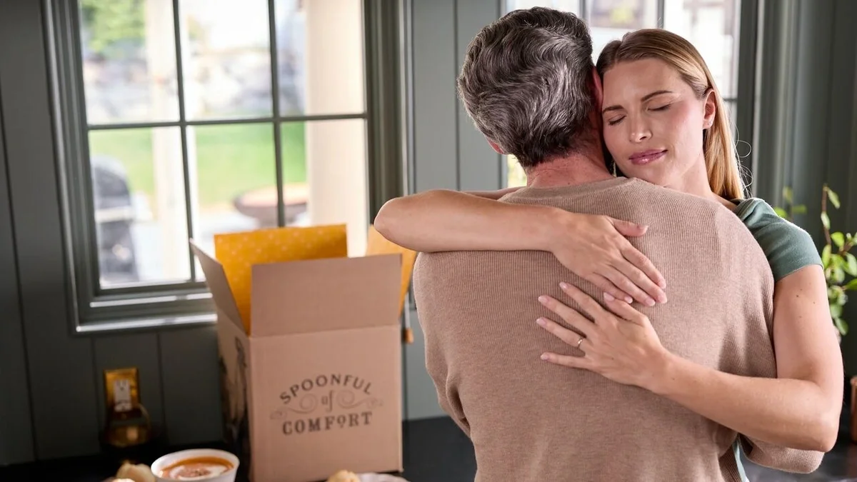 A blonde woman wearing a wedding band gingerly hugs a man with salt and pepper hair, with a Spoonful of Comfort soup package in the background.