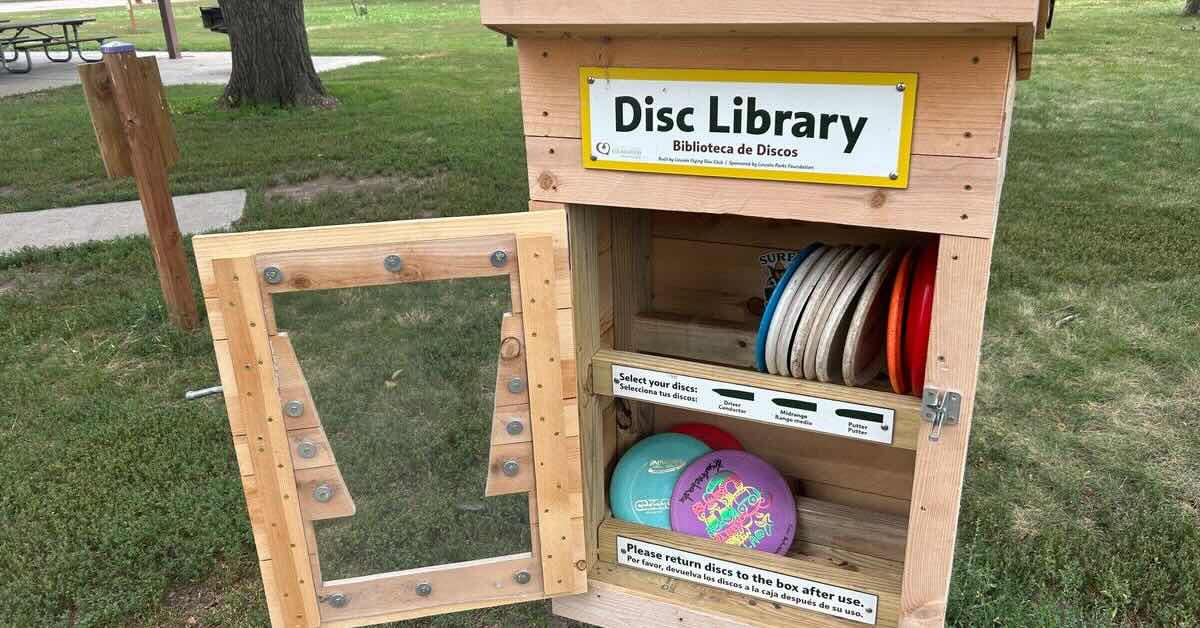A wooden cabinet in a park with disc golf discs in it and a sign saying "Disc Library"