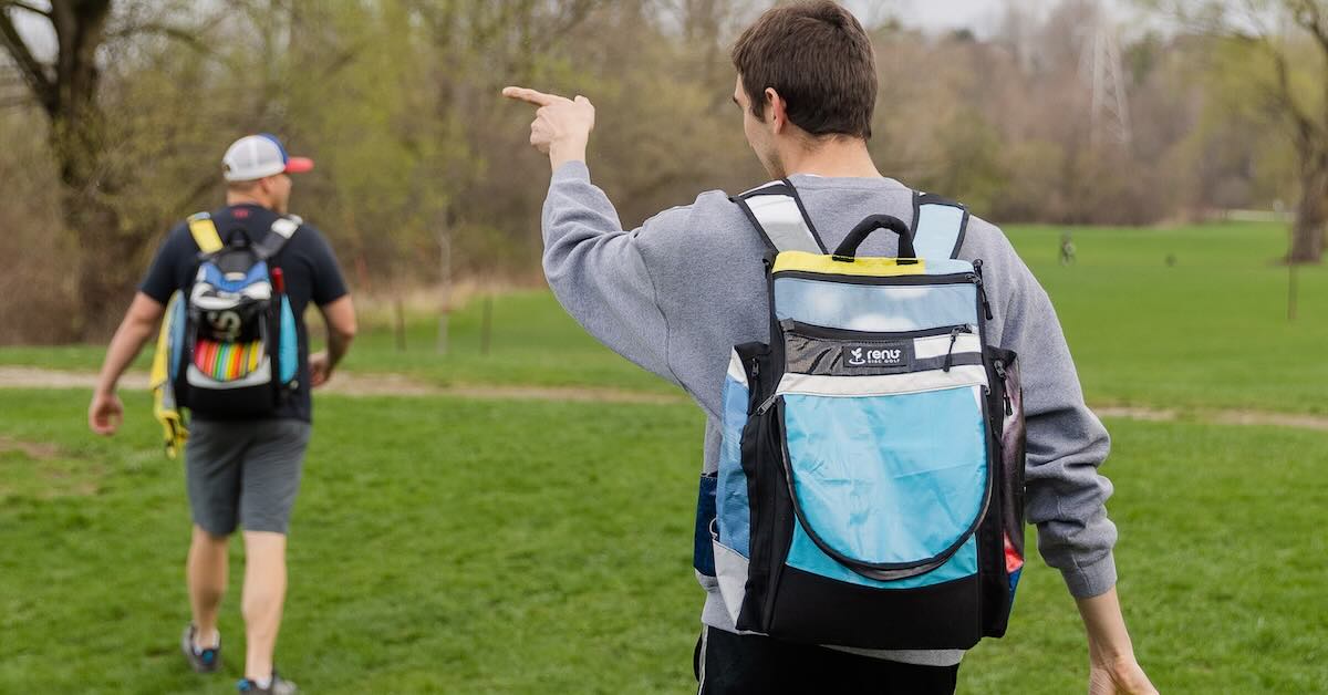 A group of disc golfers with backpacks walking on the course. In the foreground, one is pointing forward