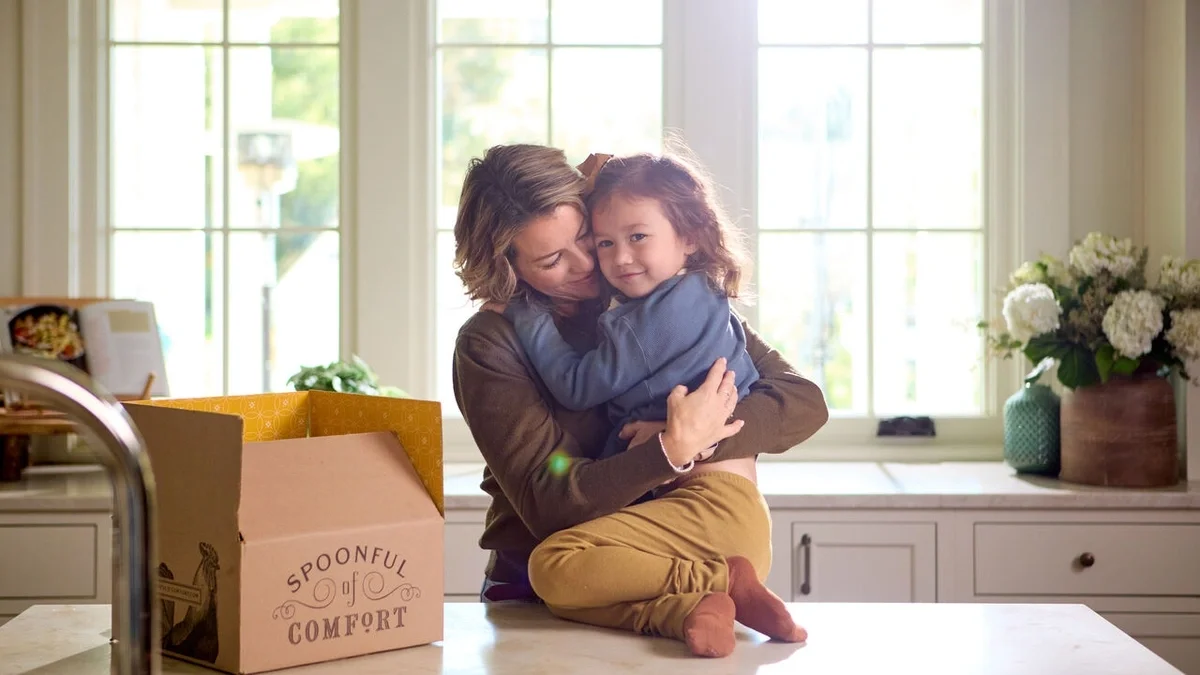 Parent standing at a kitchen counter and embracing a young child, with an open Spoonful of Comfort gift box nearby in a bright, welcoming kitchen.