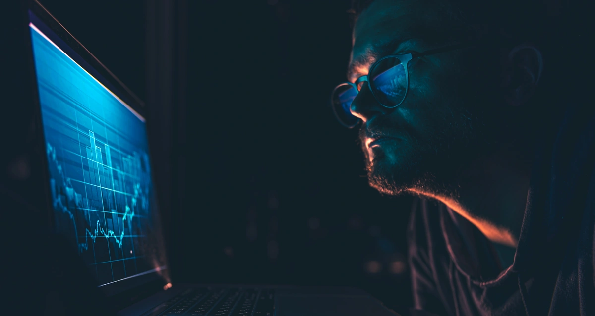 A man wearing glasses intently analyzing stock market charts and financial data displayed on a laptop screen in a dimly lit room.